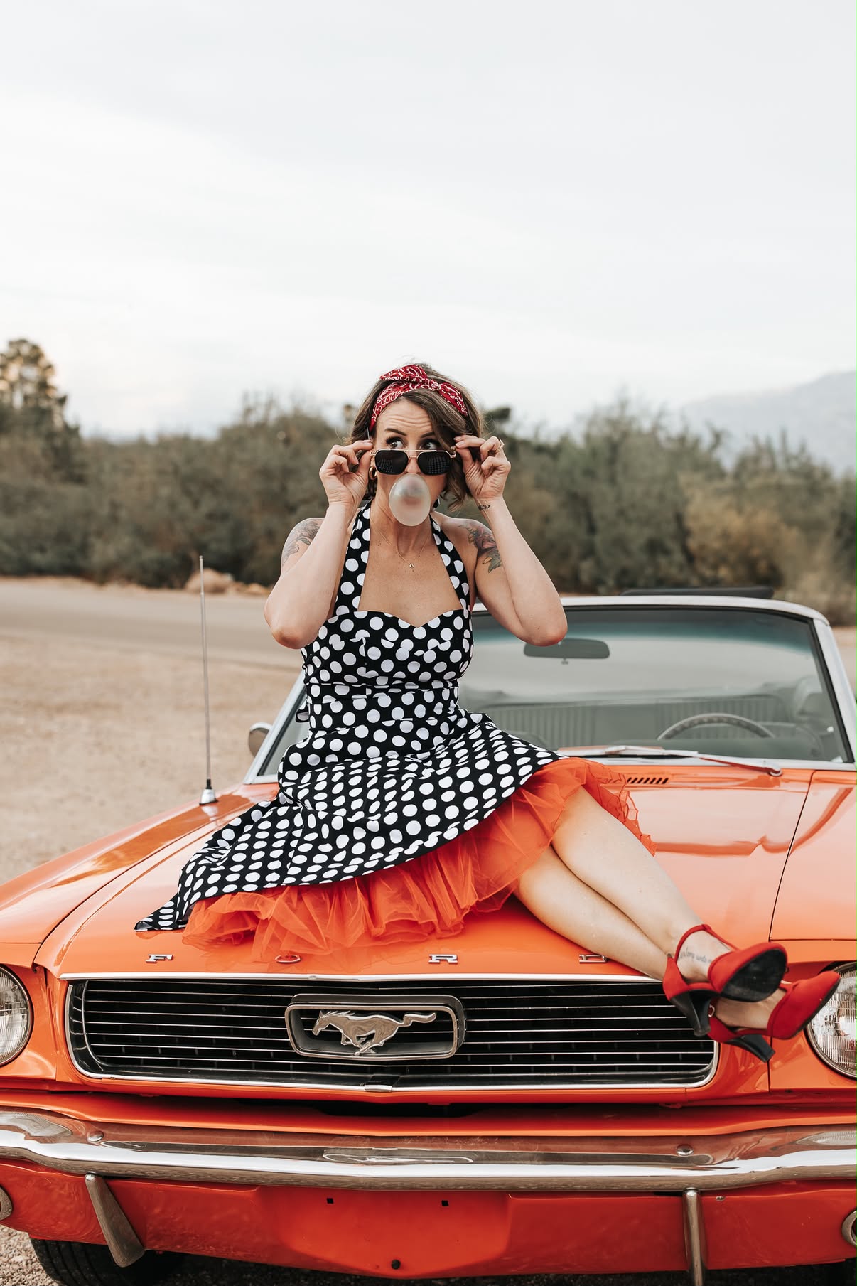 Woman in a polka-dot dress blowing bubblegum while seated on the red Mustang